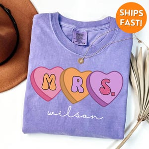 May include: A purple t-shirt with a graphic of three candy hearts that spell out "MRS." with the last heart having a period. The text "wilson" is written below the hearts.