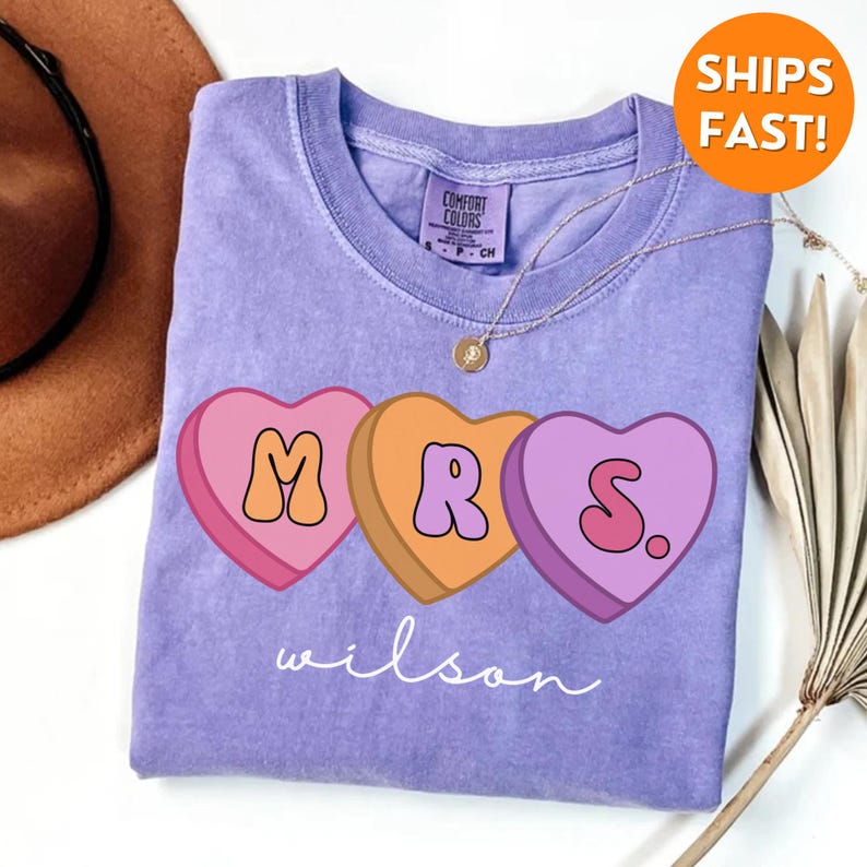 May include: A purple t-shirt with a graphic of three candy hearts that spell out "MRS." with the last heart having a period. The text "wilson" is written below the hearts.