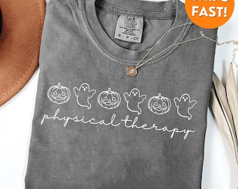 May include: A gray Comfort Colors t-shirt featuring a white graphic of pumpkins and ghosts, with the words "physical therapy" in a script font. A silver necklace with a pendant is draped across the shirt.