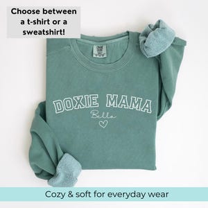 May include: A teal green sweatshirt with the text "DOXIE MAMA" in white outlined letters. The name "Bella" is written in cursive below the text with a small heart.