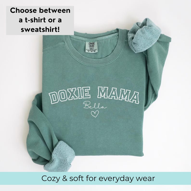 May include: A teal green sweatshirt with the text "DOXIE MAMA" in white outlined letters. The name "Bella" is written in cursive below the text with a small heart.