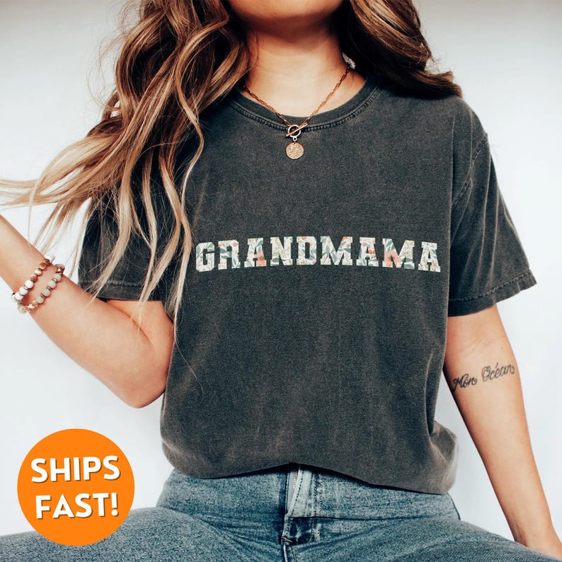 以下が含まれることがあります： 花柄の白い文字で「GRANDMAMA」とプリントされた濃いグレーのTシャツ。文字は白で縁取られています。