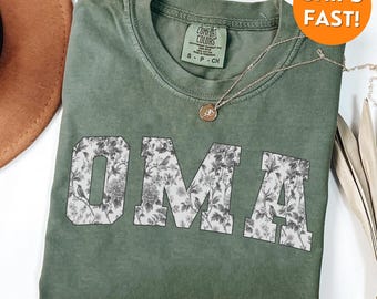 Puede incluir: Una camiseta verde oliva con la palabra "OMA" en letras grandes con estampado floral. La camiseta tiene cuello redondo y mangas cortas. Un collar dorado con un colgante está drapeado sobre la camiseta. La camiseta está hecha de un material suave y cómodo.