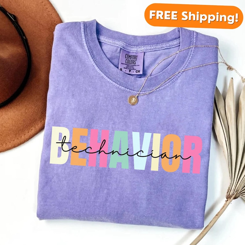 May include: A purple t-shirt with the text "BEHAVIOR technician" in rainbow colors. The text is in a playful, handwritten font.