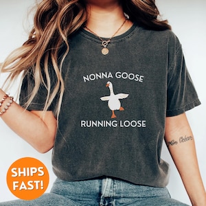 May include: Dark gray t-shirt with the text "NONNA GOOSE RUNNING LOOSE" above a cartoon goose illustration. The goose is white with orange feet and a beak, and is in a dancing pose. The shirt is a casual style.