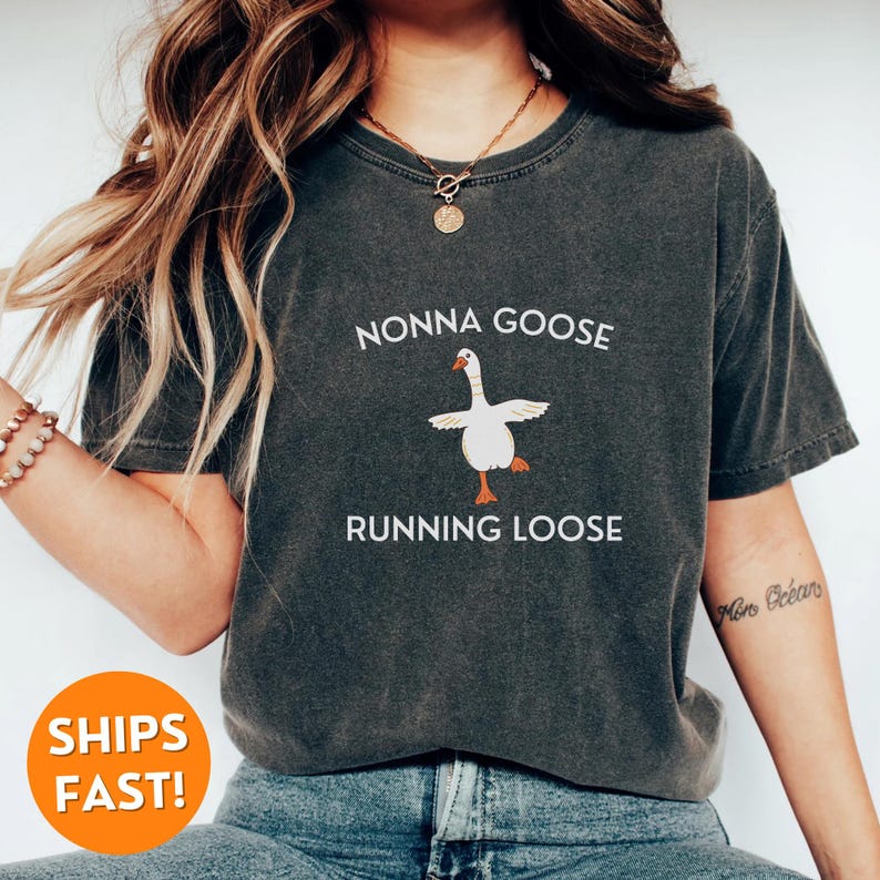 May include: Dark gray t-shirt with the text "NONNA GOOSE RUNNING LOOSE" above a cartoon goose illustration. The goose is white with orange feet and a beak, and is in a dancing pose. The shirt is a casual style.