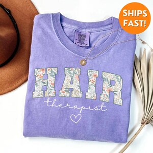 May include: Purple t-shirt with a floral design that says "HAIR therapist" with a heart below the text.