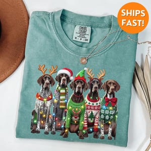 May include: A teal blue t-shirt featuring a Christmas design with four German Shorthaired Pointers wearing festive sweaters and reindeer antlers. The dogs are surrounded by Christmas lights.