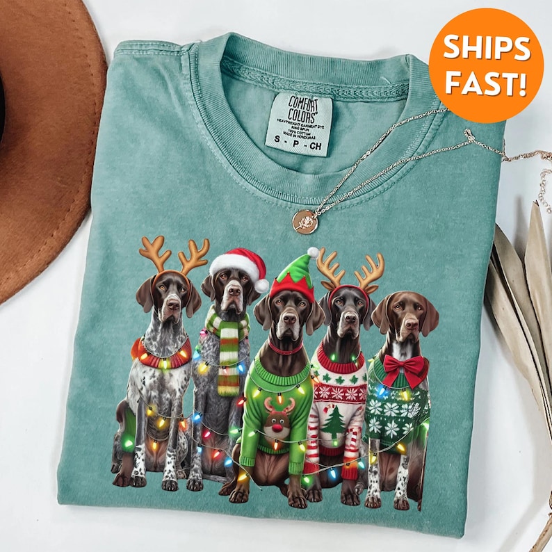 May include: A teal blue t-shirt featuring a Christmas design with four German Shorthaired Pointers wearing festive sweaters and reindeer antlers. The dogs are surrounded by Christmas lights.