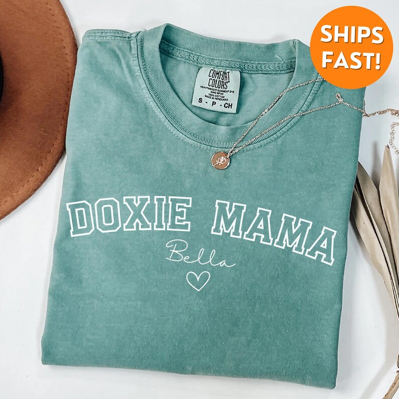 May include: A teal blue t-shirt with the text "DOXIE MAMA" in white with a white outline. The text "Bella" is written in cursive below the main text with a small heart.
