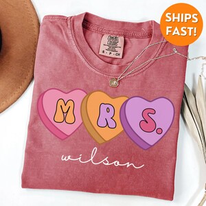 May include: A pink, orange, and purple heart-shaped graphic on a red t-shirt reads "MRS. wilson".