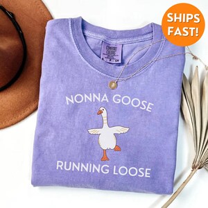 May include: A lilac-colored t-shirt with the words "NONNA GOOSE RUNNING LOOSE" in white. The shirt features a cartoon goose illustration. A gold necklace with a pendant is draped over the shirt.