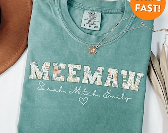 May include: A teal blue t-shirt with a floral print design that says "MEEMAW" in block letters. The names "Sarah, Mitch, Emily" are written below the word "MEEMAW" with a heart symbol.