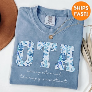 May include: A light blue t-shirt with a floral print design that spells out "OTA" for Occupational Therapy Assistant.