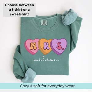 May include: A teal sweatshirt with a graphic of three candy hearts spelling "MRS." with the last heart having a period. The text "wilson" is printed below the hearts.