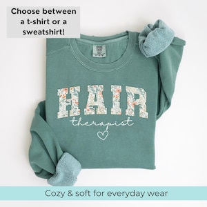 May include: A teal green sweatshirt with the text "HAIR therapist" in white with a floral pattern behind the letters. A white heart is below the text.