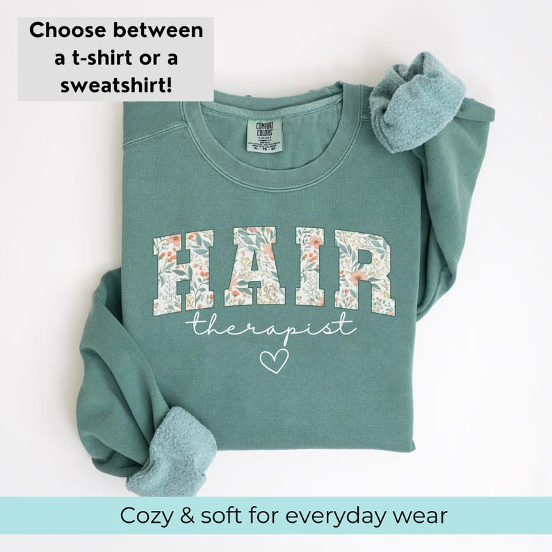 May include: A teal green sweatshirt with the text "HAIR therapist" in white with a floral pattern behind the letters. A white heart is below the text.