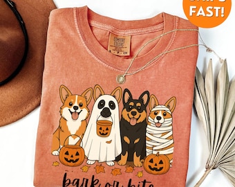 May include: Orange Comfort Colors t-shirt featuring a Halloween-themed graphic of four corgi dogs dressed as a pumpkin, ghost, vampire, and mummy. The phrase "bark or bite" is printed below the dogs. A gold necklace is draped over the shirt.