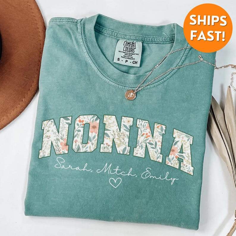 May include: A teal blue t-shirt with a floral print design that says "NONNA" in large letters. The shirt also has the names "Sarah, Mitch, Emily" and a heart printed below the floral design.
