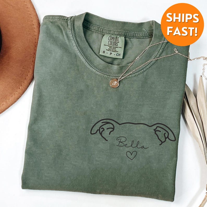 Pode incluir: Uma camiseta verde com um desenho de linha preta de uma cabe&ccedil;a de cachorro com o nome "Bella" e um cora&ccedil;&atilde;o abaixo.