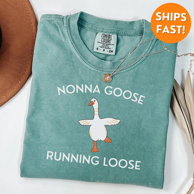 May include: A teal Comfort Colors t-shirt with the words "NONNA GOOSE RUNNING LOOSE" and a cartoon goose graphic. A silver necklace with a pendant is draped across the shirt. The shirt is a size small.