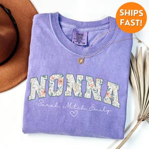 May include: Purple t-shirt with a floral design that says "NONNA" in a vintage collegiate font. The shirt also has the names "Sarah, Mitch, Emily" and a heart printed below the main design.
