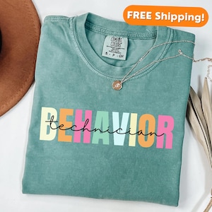 May include: A teal blue t-shirt with the text "BEHAVIOR technician" in rainbow colors. The text is in a playful, casual font.