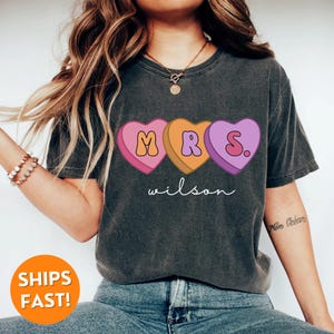 May include: A dark gray t-shirt with a graphic of three candy hearts that spell out "MRS." with the last heart having a period. The text "wilson" is printed below the hearts.