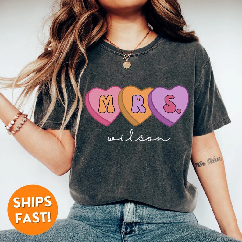 May include: A dark gray t-shirt with a graphic of three candy hearts that spell out "MRS." with the last heart having a period. The text "wilson" is printed below the hearts.