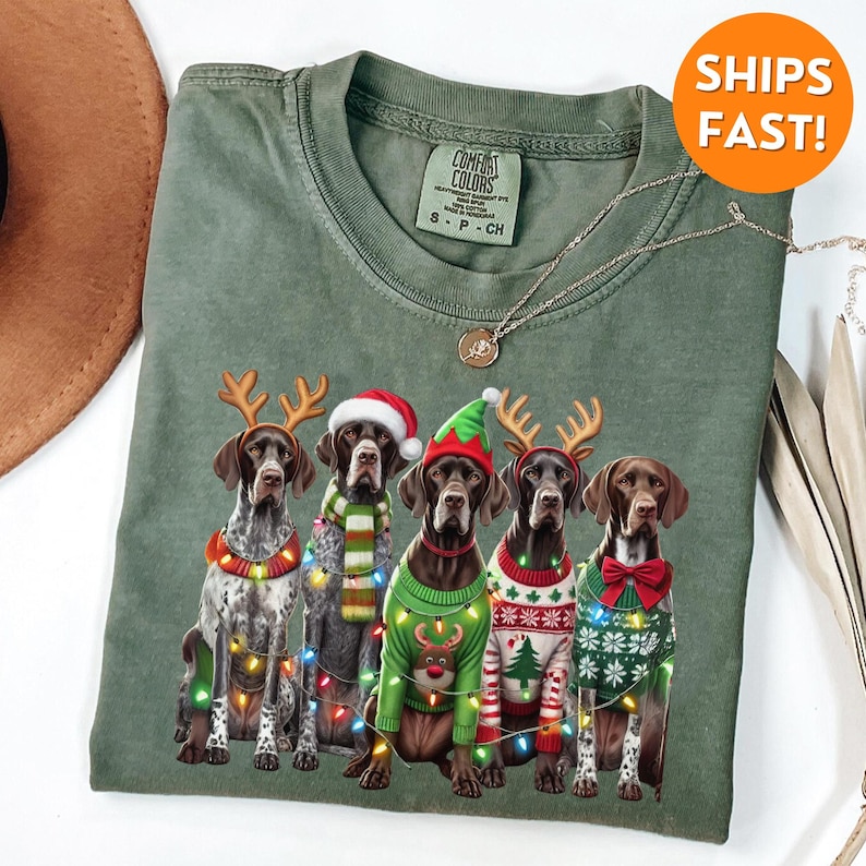 May include: Five German Shorthaired Pointers wearing Christmas jumpers and reindeer antlers with Christmas lights around them. The dogs are sitting in a row on a green background.