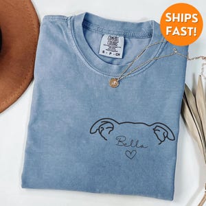 Pode incluir: Uma camiseta azul claro com um desenho de linha preta de uma cabe&ccedil;a de cachorro com o nome "Bella" e um cora&ccedil;&atilde;o abaixo.