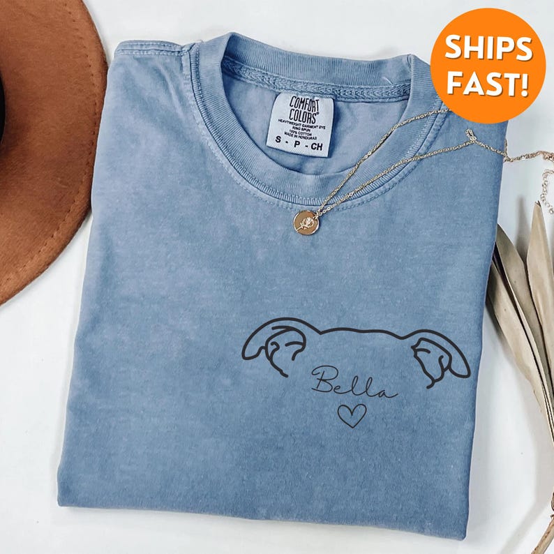 Pode incluir: Uma camiseta azul claro com um desenho de linha preta de uma cabe&ccedil;a de cachorro com o nome "Bella" e um cora&ccedil;&atilde;o abaixo.