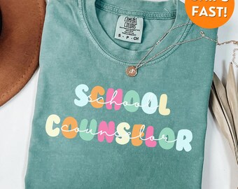 May include: A teal Comfort Colors t-shirt with the words "School Counselor" in colorful bubble letters. A silver necklace with a pendant is draped on the shirt. The shirt is a size small.