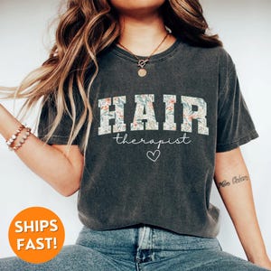 May include: A dark gray t-shirt with a floral print design that says "HAIR therapist" with a white heart underneath.