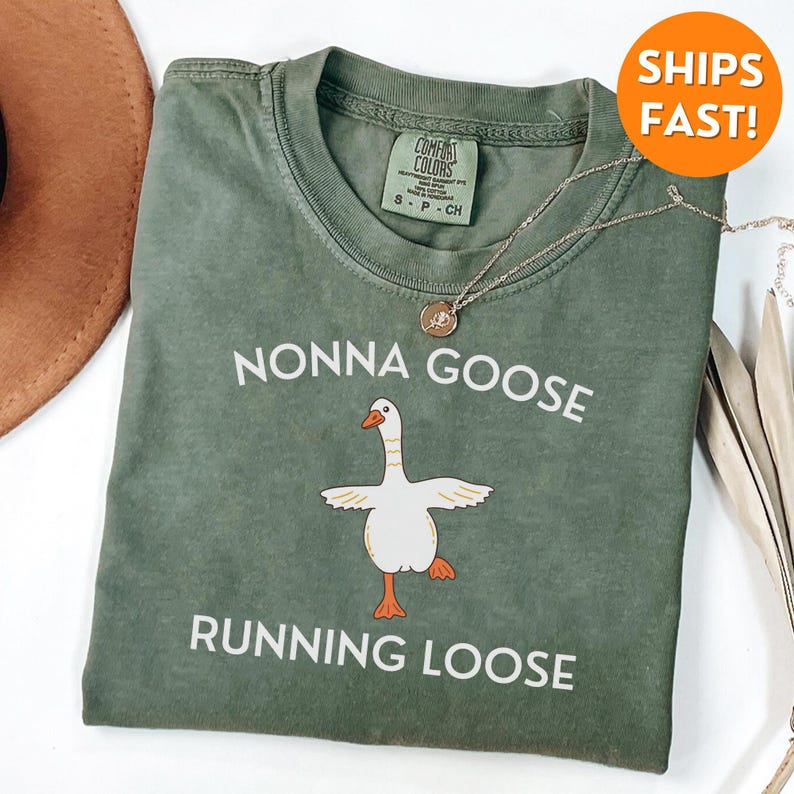 May include: A sage green t-shirt with the words "NONNA GOOSE RUNNING LOOSE" in white. The shirt features a cartoon goose illustration with outstretched wings and orange feet. A silver necklace with a pendant is draped across the shirt.