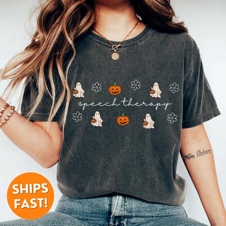 May include: Dark gray t-shirt with the words "speech therapy" in a cursive font. The shirt features a pattern of pumpkins, ghosts, and flowers. The shirt is made of a soft material.