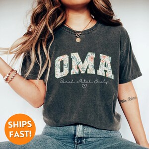 May include: A dark grey t-shirt with a floral print design that spells out "OMA" in large letters. The shirt also has the text "Sarah, Mitch, Emily" and a small heart printed below the floral design.