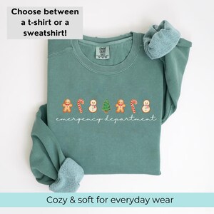 Puede incluir: Una sudadera de color verde azulado con un dise&ntilde;o navide&ntilde;o. El dise&ntilde;o presenta hombres de jengibre, bastones de caramelo, mu&ntilde;ecos de nieve y &aacute;rboles de Navidad. El texto "emergency department" est&aacute; escrito debajo del dise&ntilde;o.