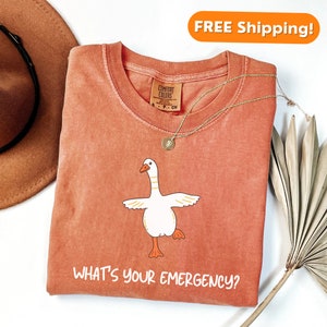 May include: A peach-colored t-shirt with a white goose illustration and the text "What's your emergency?" printed on the front. The shirt is folded and laid flat on a white surface.