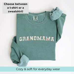 以下が含まれることがあります： 花柄の文字で「GRANDMAMA」とプリントされたティールグリーンのスウェットシャツ。スウェットシャツは折り畳まれ、袖はまくり上げられています。