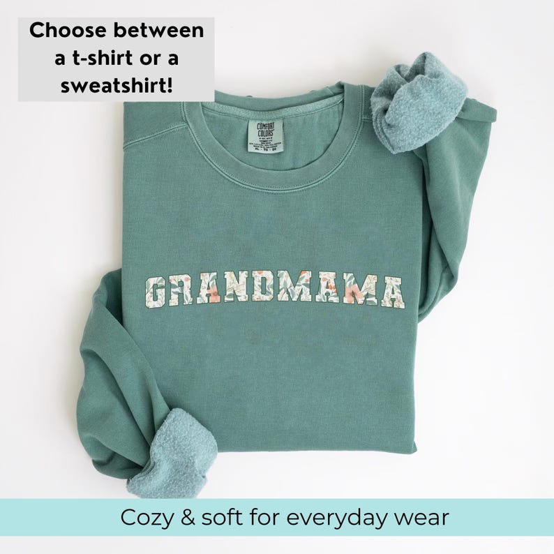 以下が含まれることがあります： 花柄の文字で「GRANDMAMA」とプリントされたティールグリーンのスウェットシャツ。スウェットシャツは折り畳まれ、袖はまくり上げられています。