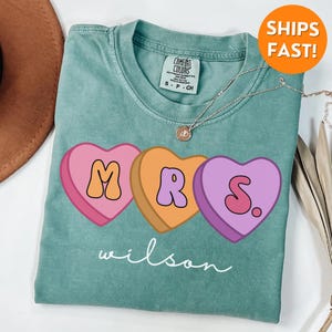 May include: A teal blue t-shirt with a graphic of three candy hearts that spell out "MRS." with the last heart having a period after the "S". The text "wilson" is written in cursive below the hearts.