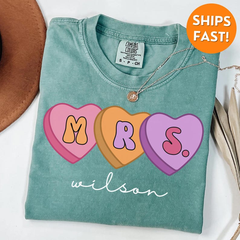 May include: A teal blue t-shirt with a graphic of three candy hearts that spell out "MRS." with the last heart having a period after the "S". The text "wilson" is written in cursive below the hearts.