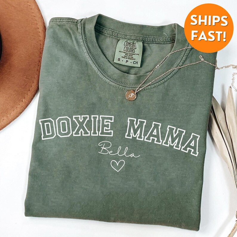 May include: Green t-shirt with the text "Doxie Mama" in white with a white outline. The text "Bella" is written in cursive below the main text with a small heart.