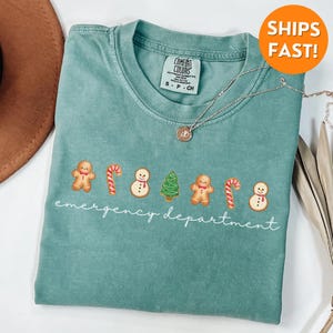 Puede incluir: Una camiseta azul verdosa con un gr&aacute;fico blanco de hombres de jengibre, bastones de caramelo, mu&ntilde;ecos de nieve y &aacute;rboles de Navidad. El texto "emergency department" est&aacute; escrito debajo del gr&aacute;fico.