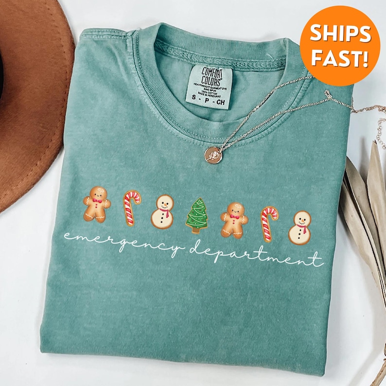 Puede incluir: Una camiseta azul verdosa con un gr&aacute;fico blanco de hombres de jengibre, bastones de caramelo, mu&ntilde;ecos de nieve y &aacute;rboles de Navidad. El texto "emergency department" est&aacute; escrito debajo del gr&aacute;fico.