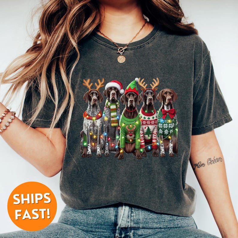 May include: A dark grey t-shirt featuring a graphic of five German Shorthaired Pointers wearing Christmas jumpers and reindeer antlers. The dogs are surrounded by Christmas lights.