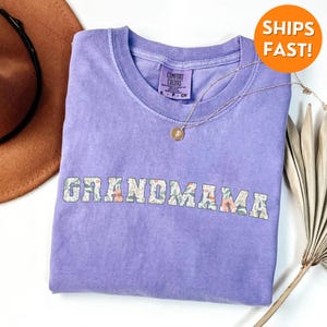 以下が含まれることがあります： 花柄の白い文字で「GRANDMAMA」とプリントされた紫色のTシャツ。柔らかく快適な生地で作られています。
