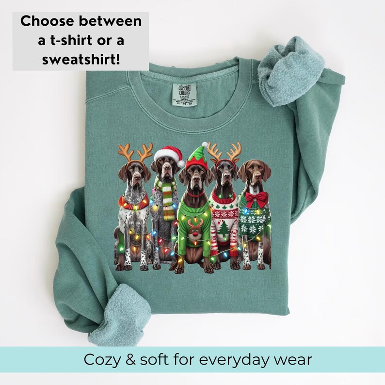 May include: A teal green sweatshirt featuring a Christmas design with five German Shorthaired Pointers wearing festive sweaters and reindeer antlers. The dogs are surrounded by Christmas lights.
