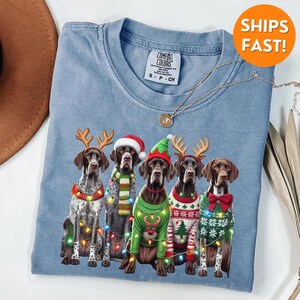 May include: A blue t-shirt with a Christmas-themed design featuring five German Shorthaired Pointers wearing reindeer antlers and festive sweaters with Christmas lights. The text "COMFORT COLORS" is visible on the tag.